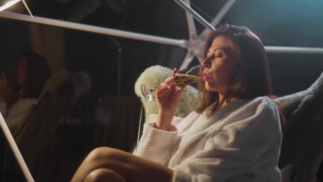 disappointed lady drinks champagne at midnight on transparent terrace. woman relieves stress with alcoholic beverage sitting in furry armchair by large window