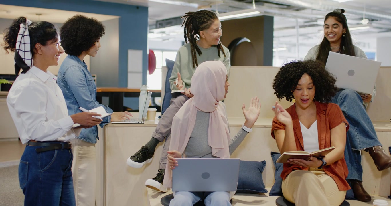 Diverse team watching woman in pink scarf opening meeting at office, gesturing with laptop