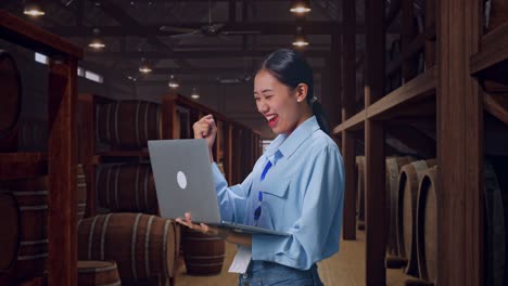 side view of an asian female professional worker standing with her laptop in cellar of winery, she raises her fist up with screaming goal after typed on the laptop