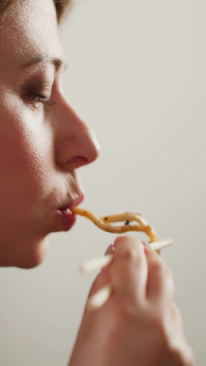 Close up of woman eating noodles with chopsticks focusing on her profile as woman brings food to mouth. Soft lighting highlights facial features and texture details against plain background