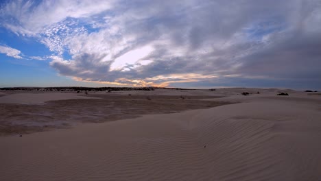 A-setting-sun-tints-the-clouds-orange-and-pink,-as-they-pass-over-the-sand-dunes-at-Gran-Desierto-de-Altar-Biosphere-Reserve,-also-known-as-El-Guapo,-Mexico