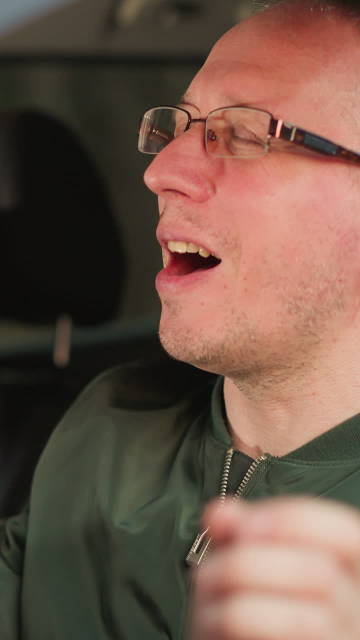 한 남자가 녹색 재과 안경을 입고 노래하는 동안 음악에 미소 짓고 부드럽게 춤을 추며 차의 운전석에 앉아 있습니다.