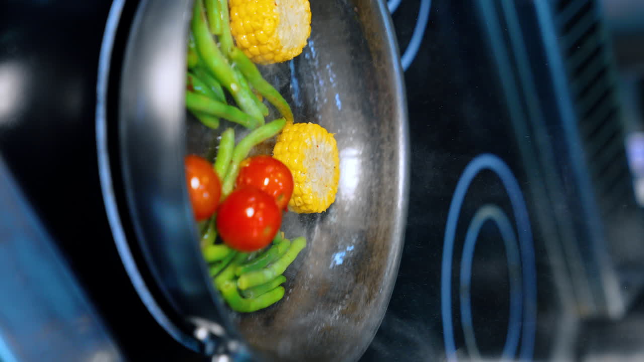 Maize and beans are fried on the frying pan. Hand in black glove throws a tomato and mixes the veggies. Close up. Vertical view.