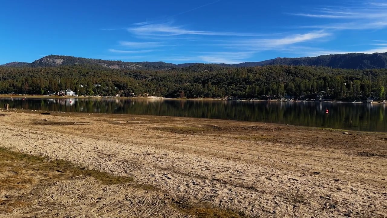 Panoramica da sinistra a destra dei bassi livelli dell'acqua al Lago Bass, California, che rivela un'ampia costa esposta e un letto del lago asciutto sullo sfondo del paesaggio montano boscoso della Sierra Nevada.