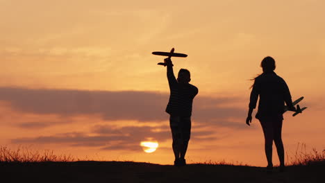 Silhouetten-Eines-Mädchens-Und-Eines-Jungen,-Die-Bei-Sonnenuntergang-Mit-Flugzeugen-Zusammen-Spielen,-Ein-Glückliches-Und-Sorgloses-Kind