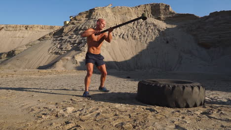 muscle athlete strongman man hits a hammer on a huge wheel in the sandy mountains in slow motion at sunset. the dust from the wheels rises.