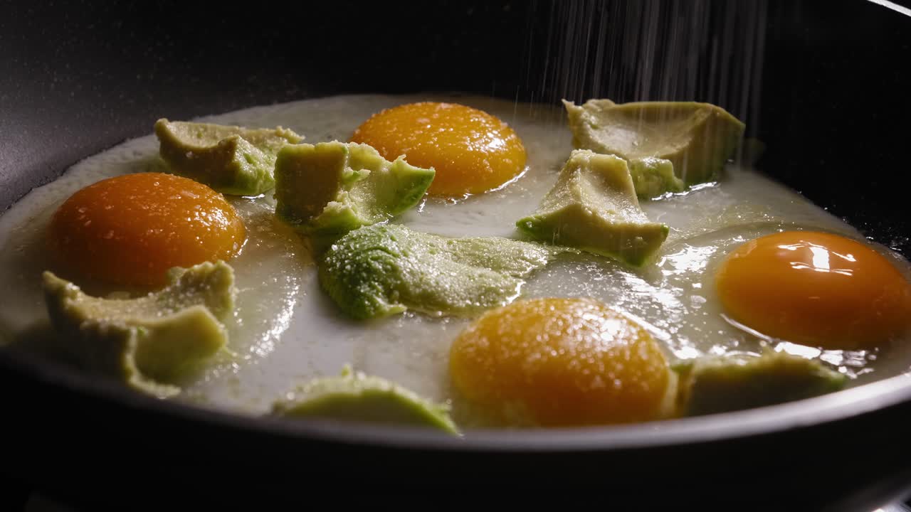 주방장이 소금과 파프리카 조미료를 부드럽게 첨가하면서 프라이팬에 익힌 날달걀과 아보카도 요리의 클로즈업 매크로 샷, 재료는 맛있는 오픈 샌드위치를 만드는 데 사용됩니다.