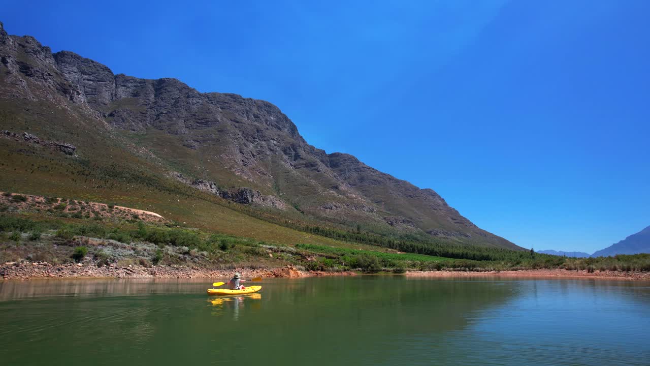 A person rowing a yellow canoe from left to right across calm turquoise waters, surrounded by dramatic mountains in South Africa’s wine region. The still pond reflects the peaks and blue sky