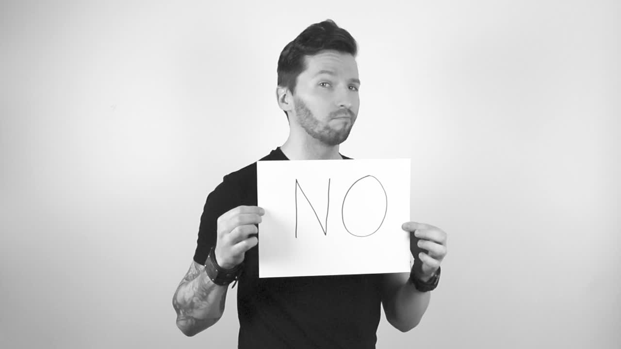 Grayscale footage of a tattooed man with short hair and beard lifting up a white paper or a sign that says NO in capital letters, man is shaking his head in denial and is not happy about something.