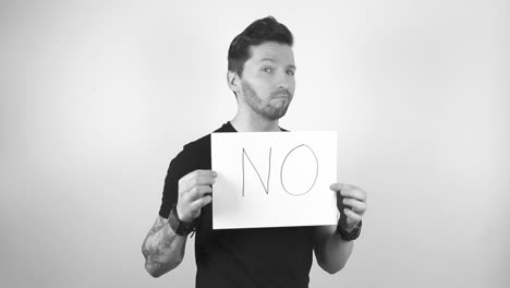 Grayscale footage of a tattooed man with short hair and beard lifting up a white paper or a sign that says NO in capital letters, man is shaking his head in denial and is not happy about something.