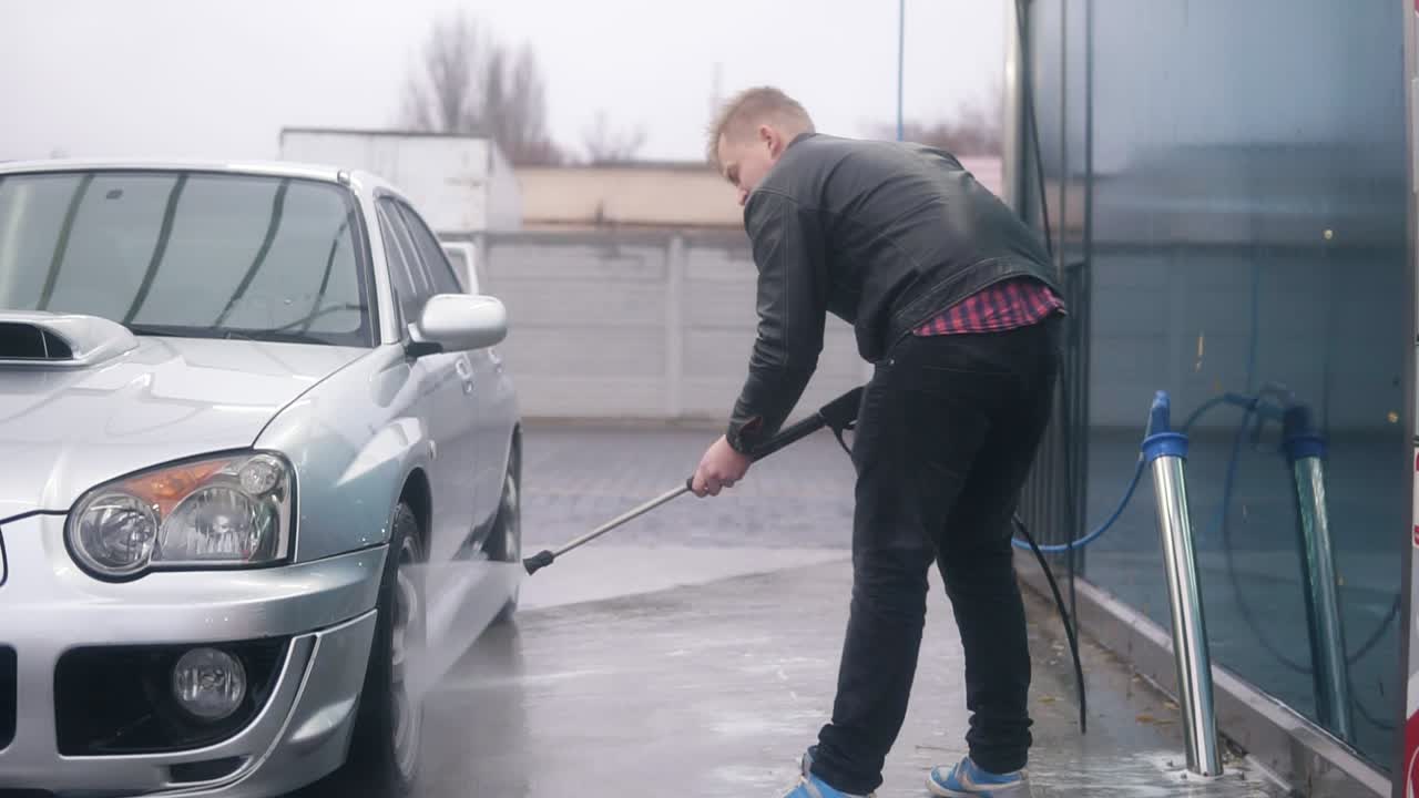 joven atractivo hombre elegante lavando su coche deportivo plateado con chorro de agua en el lavado de coches de autoservicio. él está limpiando cuidadosamente los neumáticos del coche