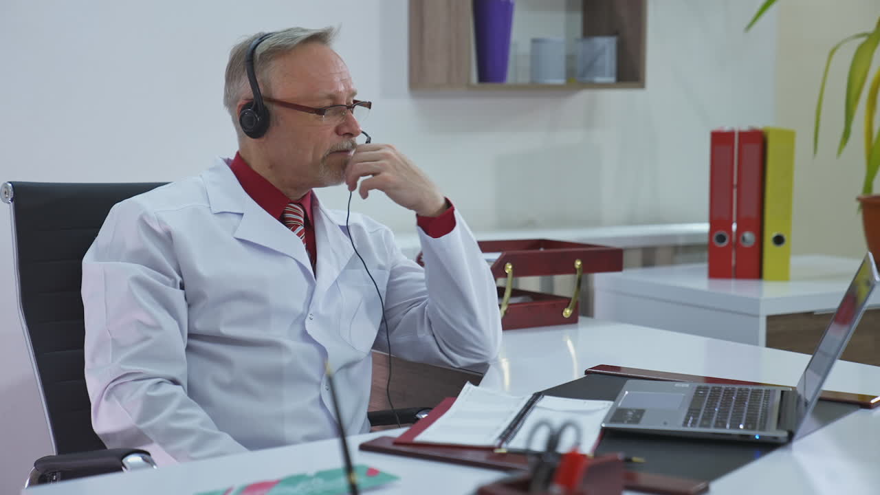 Old doctor in medical office working online. Professional male doctor in glasses and medical coat giving a consultation in front of laptop. Telemedicine. Coronavirus.