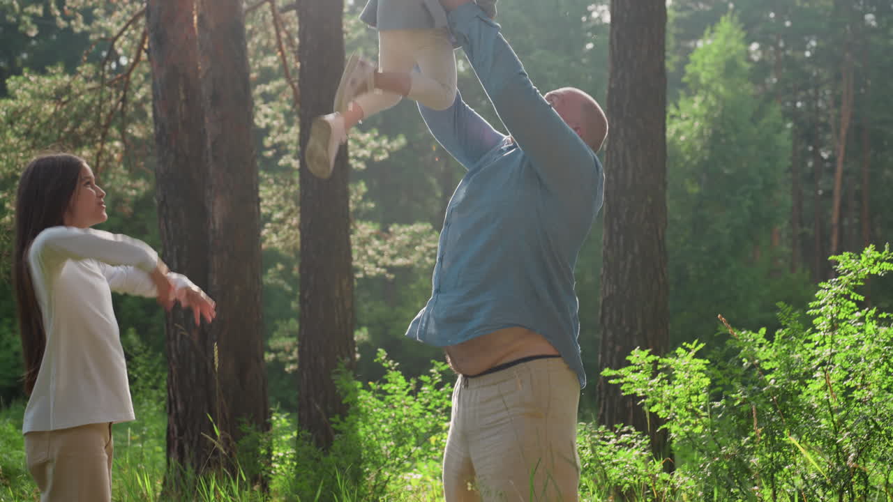Father joyfully throwing daughter up and catching her while sister watches with excitement, surrounded by lush green forest and warm sunlight, capturing fun playful family bonding
