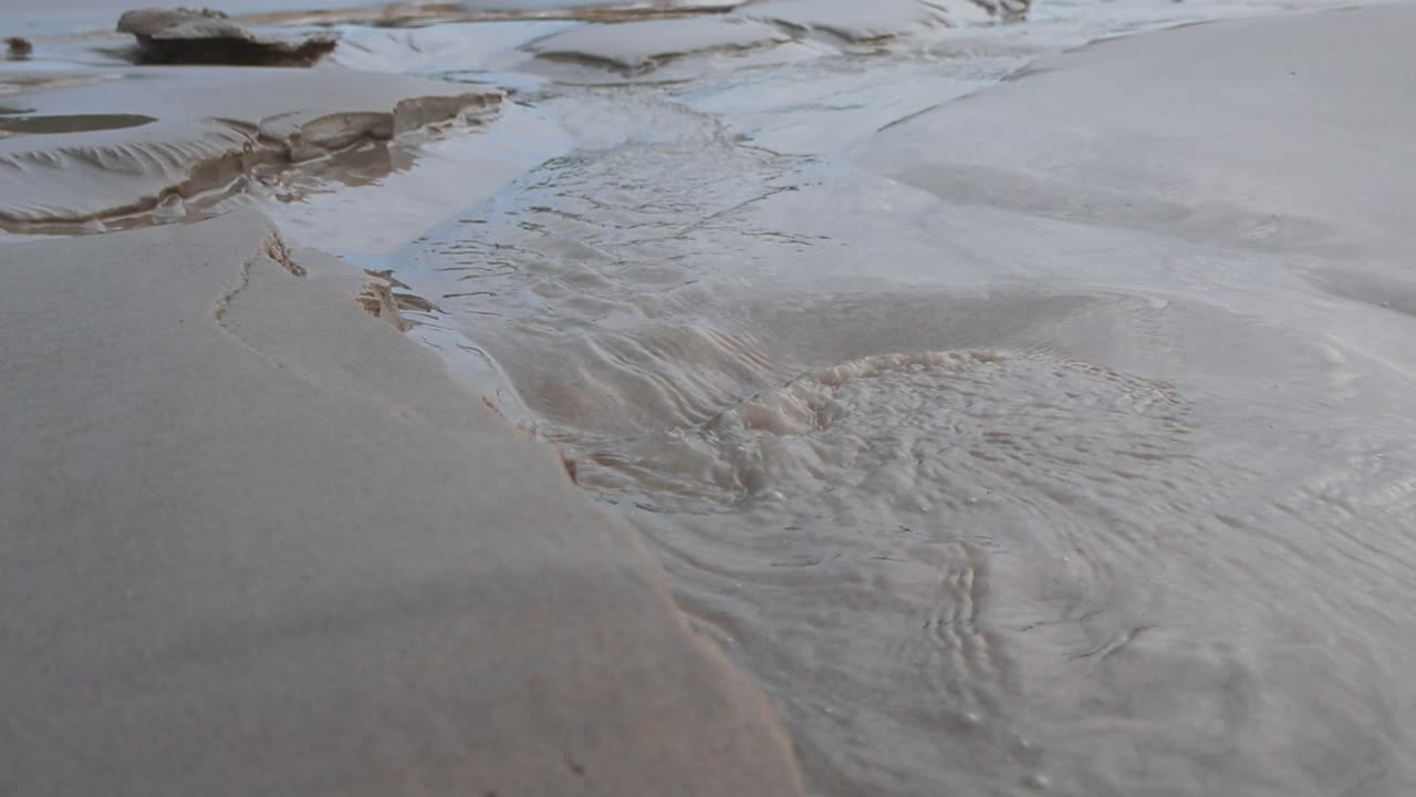 Close up or closeup footage of cold see through clear water flowing on a sandy clear beach in sharp focus and slow motion. The water is creating a pathway and has small ripples and waves, bubbles.