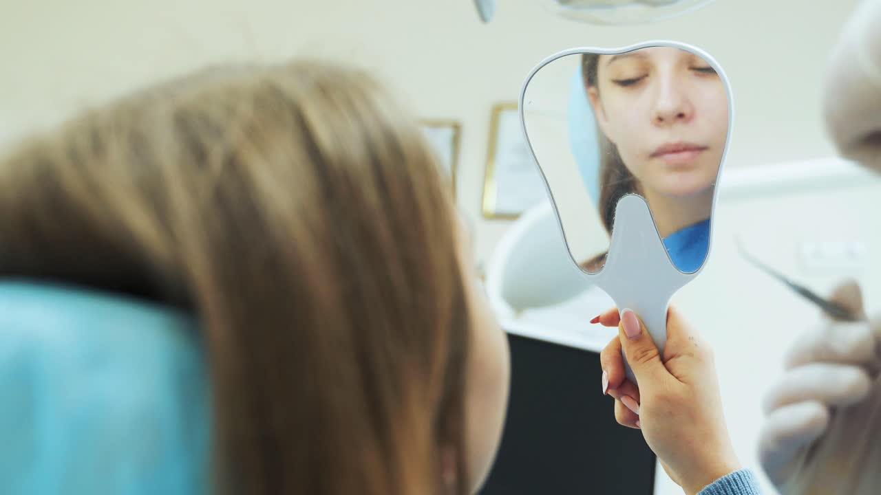 A young girl looks in the mirror in her hands as a dentist treats teeth in a clinic