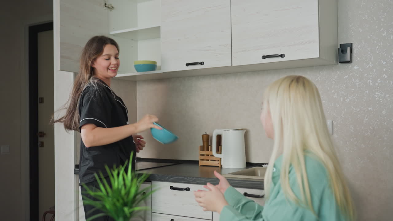 young sibling handing bowls to friend seated at kitchen table in modern apartment kitchen with light decor, fridge, microwave, and decorative plant visible, preparing breakfast together