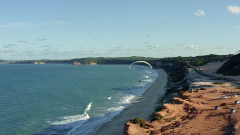 Rotating-aerial-drone-shot-of-a-paraglider-flying-over-the-stunning-tropical-northeastern-Brazil-coastline-near-Pipa-in-Rio-Grande-do-Norte-with-large-sand-dunes,-cliffs-over-the-ocean-and-tourists