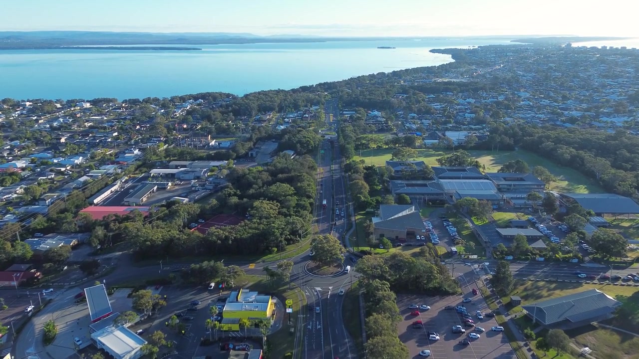 Paesaggio aereo ripreso da drone di auto che guidano attraverso una rotonda lungo una strada principale circondata da strade residenziali e abitazioni suburbane con i corsi d'acqua del lago Tuggerah, Bateau Bay, Central Coast, NSW, Australia