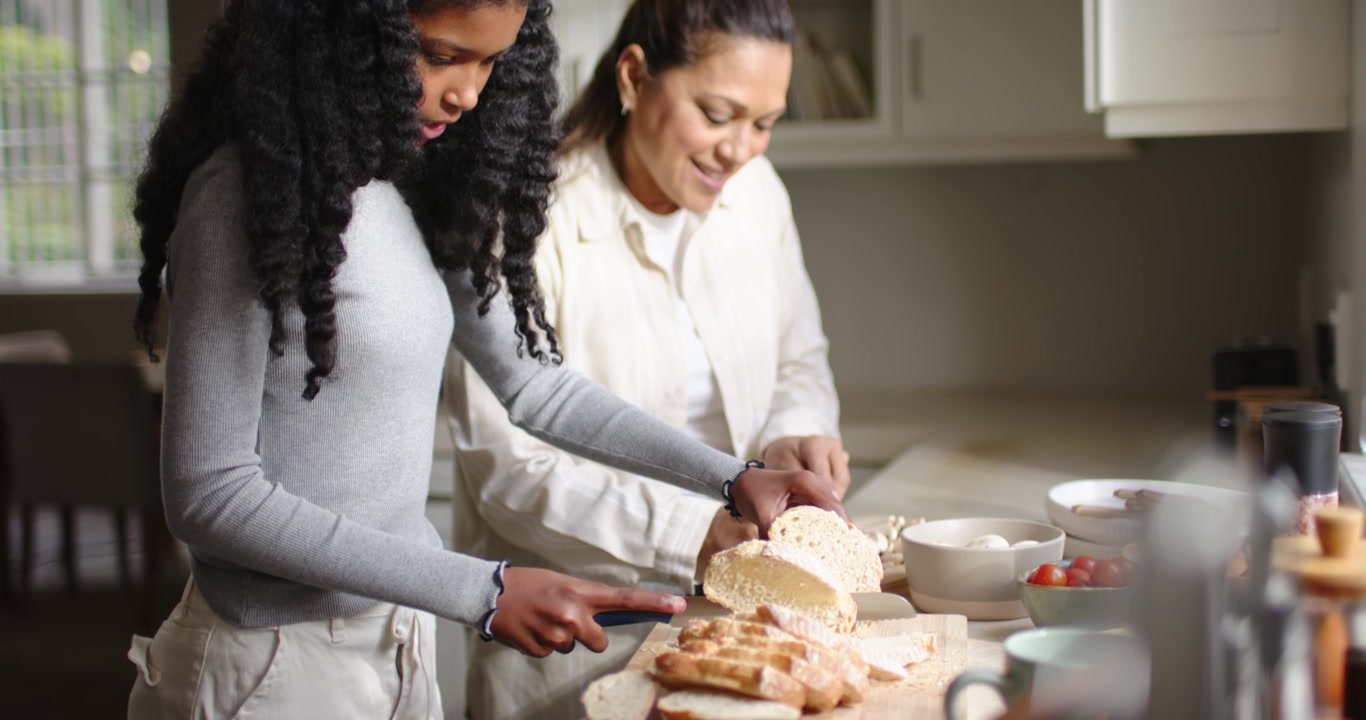 Mother and African-American teen slicing bread and cheese with knife on board making lunch at home