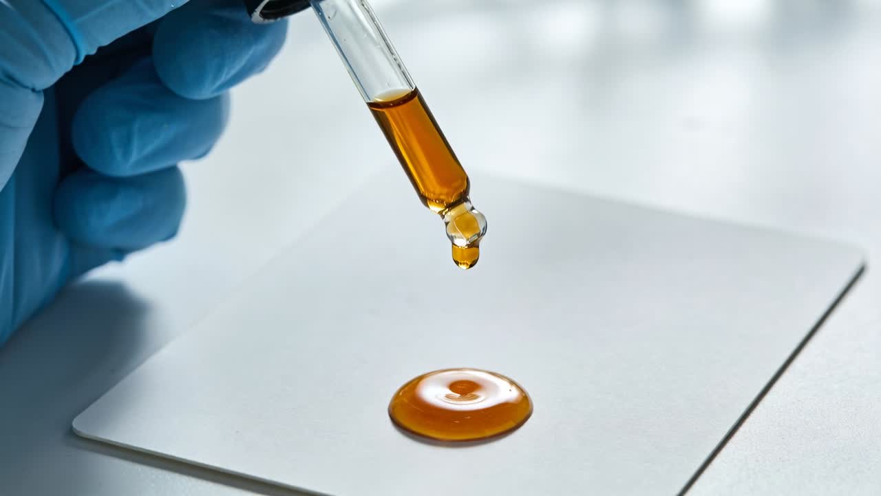 Mano guantata in nitrile blu che eroga liquido ambrato tramite contagocce su un vetrino bianco, formando una piccola pozza.