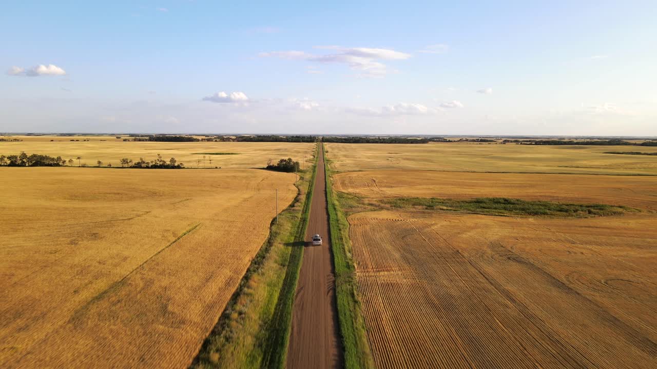 Silver car driving along straight endless dirt road swirling up dust during sunset in Canada's countryside