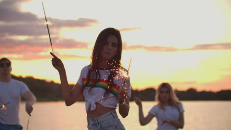 The-young-female-student-with-nude-waist-is-moving-her-hands-with-big-bengal-lights-on-the-sand-coast-with-her-friends.-This-is-theamy-summer-evening-on-the-open-air-party.