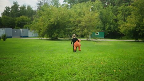 back view of young boy holding large orange teddy bear walking across lush green field surrounded by tall trees and foliage, with distant official looking structure partially visible in natural daylight setting
