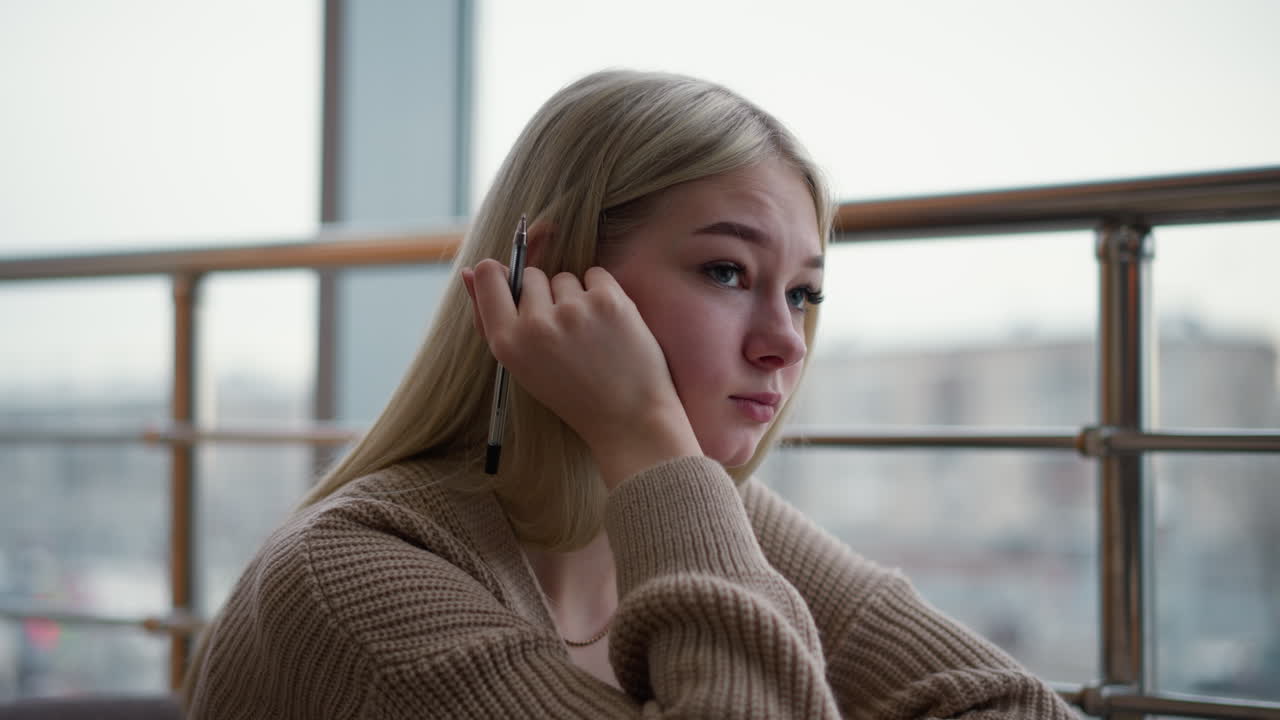 дама сидит с ручкой в руке, помещенной на подбородок, выглядит скучно, глядя на оживленную городскую обстановку через окно, мягкое размытие городской жизни снаружи