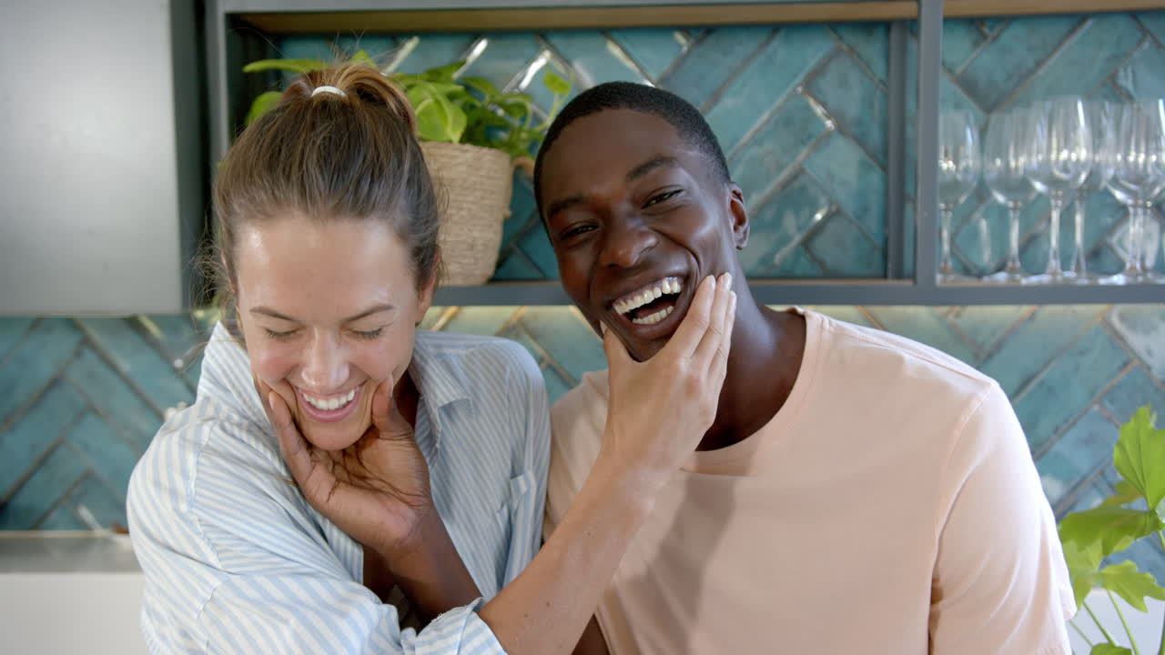 Diverse couple laughing, standing in kitchen, at home
