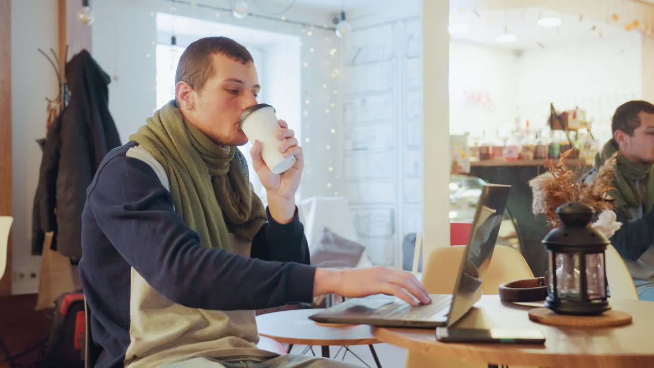 Uomo caucasico che lavora in un caffè con laptop e caffè, indossa una sciarpa verde, concentrato sullo schermo, digita sul laptop con un taccuino nelle vicinanze, luce naturale calda dalla finestra, barista sfocato sullo sfondo, atmosfera di lavoro da remoto