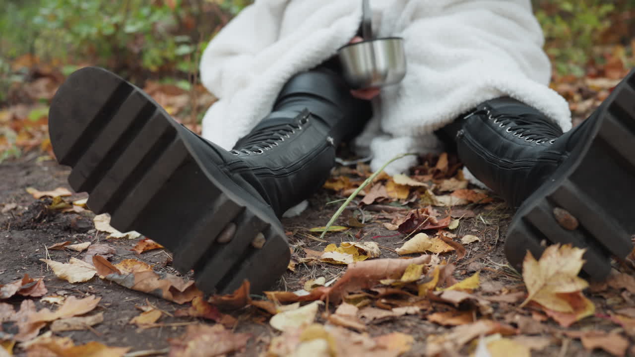 closeup sepatu bot hitam dan cangkir panas di tanah yang bertaburan daun, wanita berkemah yang terbungkus selimut menyesap minuman hangat, uap mengepul ke udara musim gugur yang sejuk, jeda perkemahan sederhana dengan cahaya alami yang lembut