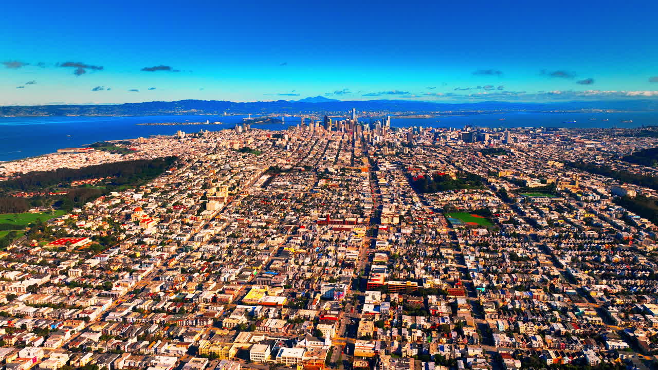Vista aerea della griglia urbana di San Francisco con lo sguardo rivolto verso il quartiere finanziario del centro. Densi edifici residenziali e commerciali si estendono dal primo piano fino alla baia. Cielo azzurro e limpido sopra.