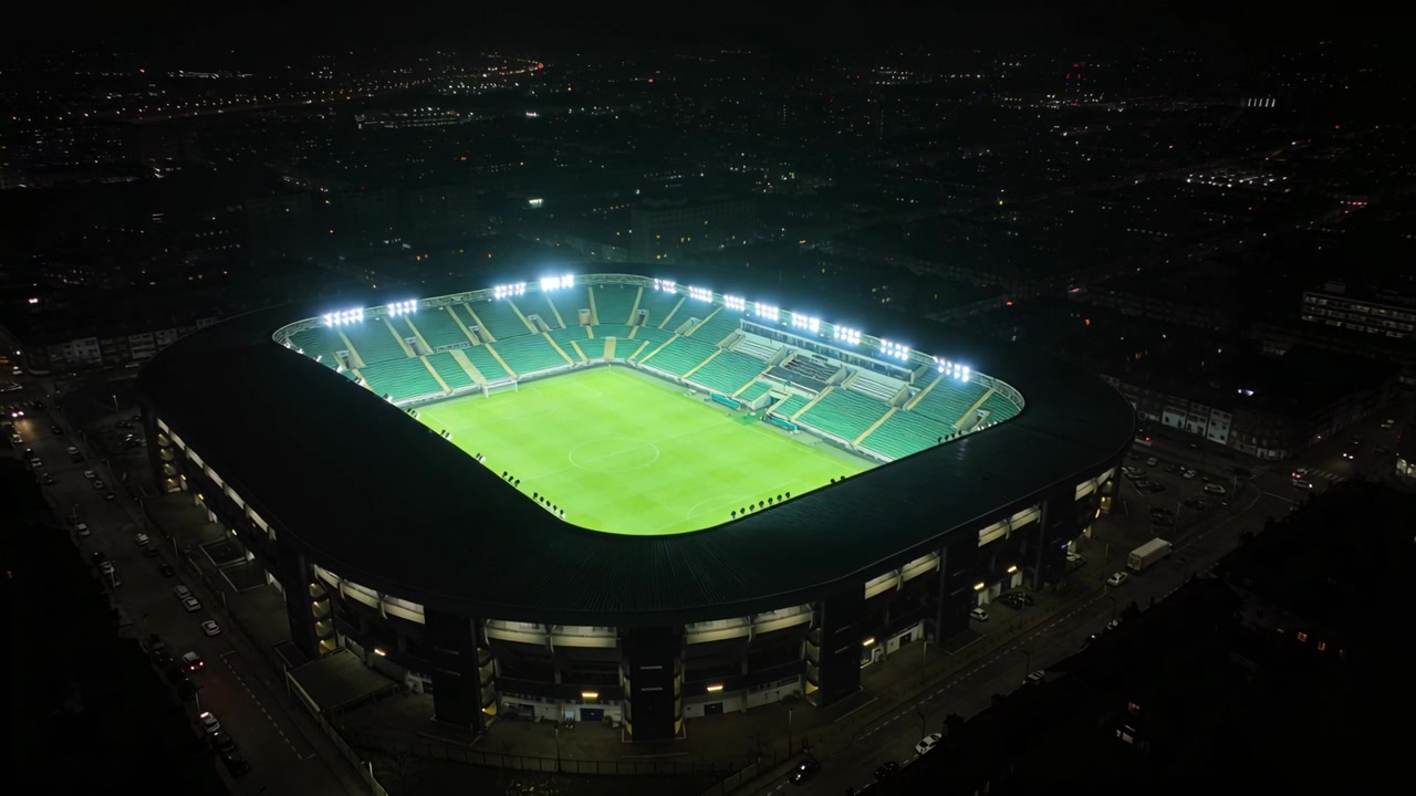 Drone in avvicinamento che si inclina e ruota sopra la conca illuminata dello stadio di notte, riprendendo il campo vuoto