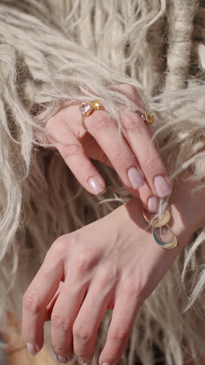 A close-up of woman's hand adorned with elegant gold rings and a unique bracelet featuring intertwined metal loops in blue and gold, set against a textured beige woolen garment