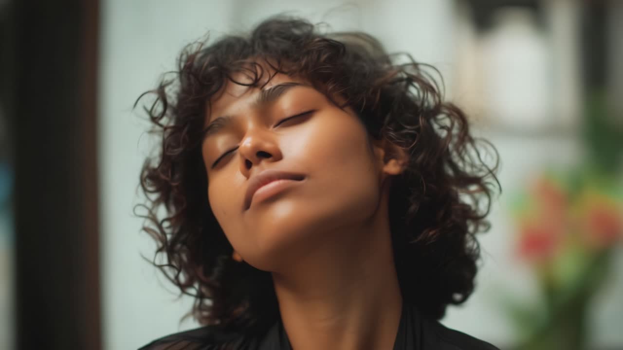 A serene woman with curly hair closes her eyes, embracing a moment of tranquility and peace while surrounded by a softly blurred, natural background