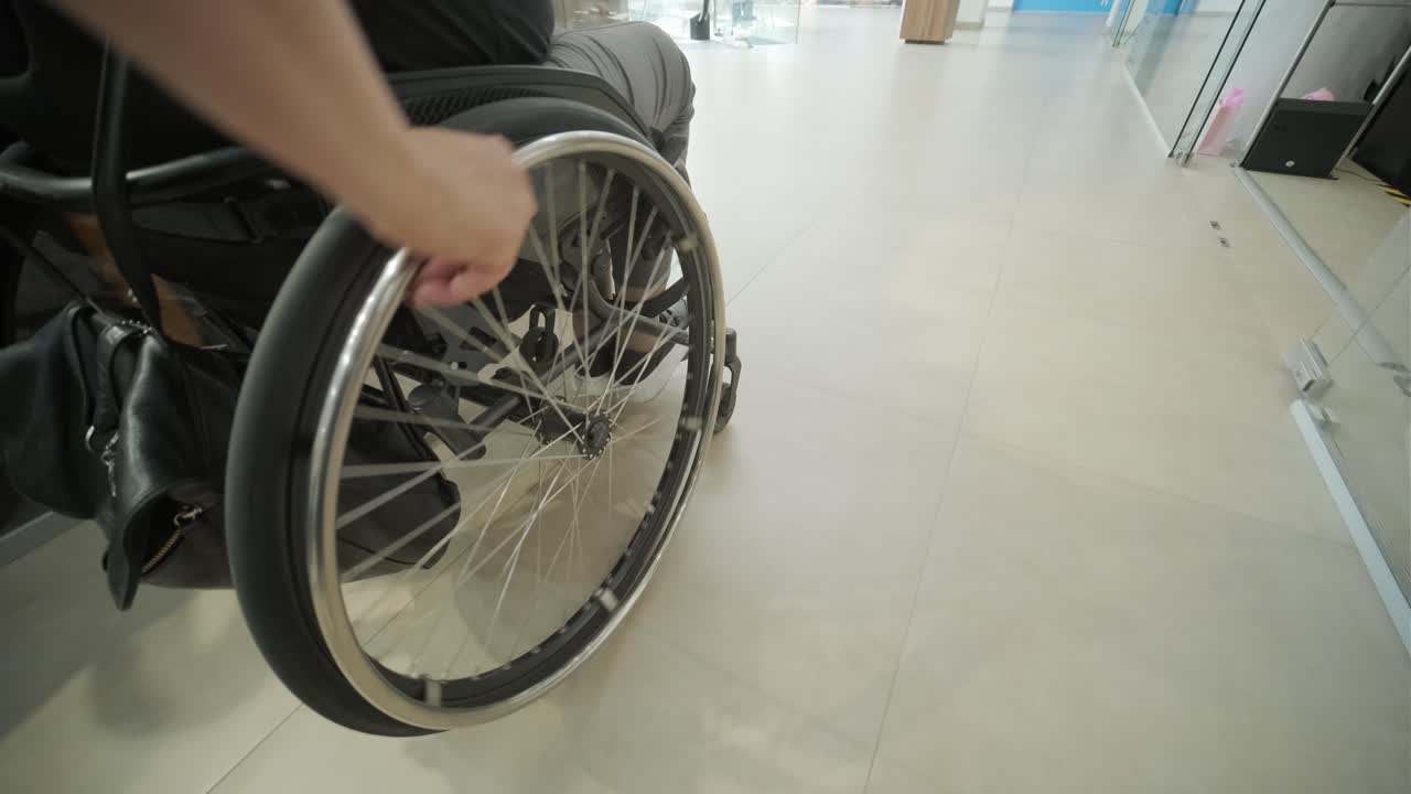 un primer plano de una persona que usa una silla de ruedas en el interior, centrándose en la rueda y la mano mientras navegan por una superficie lisa y accesible. la imagen destaca la movilidad, la independencia y el diseño accesible en espacios públicos.