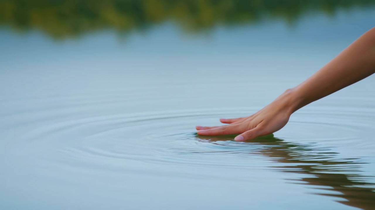 Mano che sfiora l'acqua calma del lago, leggere increspature e luce serale soffusa, primo piano della punta del dito che accarezza una superficie riflettente.