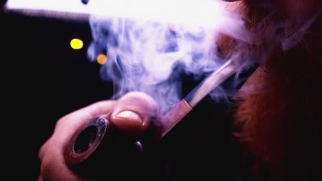 backlit man with red bushy beard smokes pipe in dark close up view