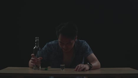 drunk asian man pouring vodka in a shot glass before drinking and sleeping in black background