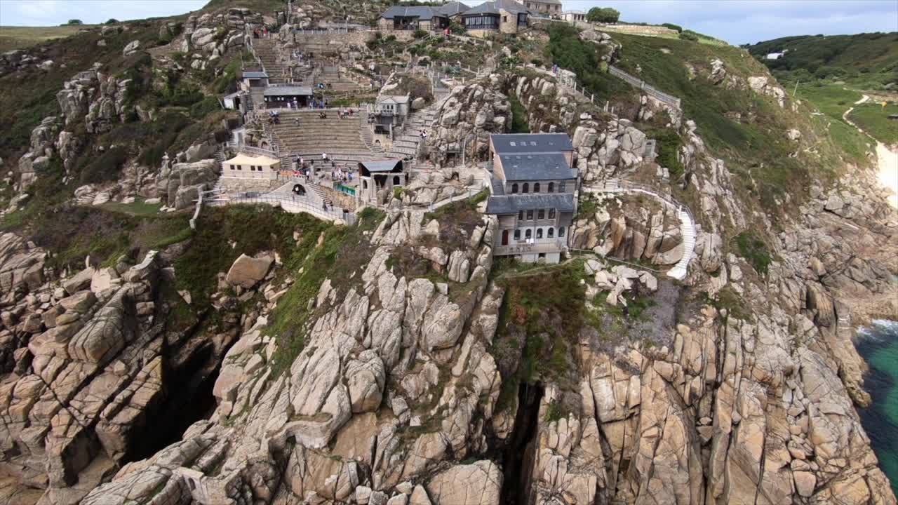 Tracking drone shot of Penzance coast line featuring the Minack Theatre and an empty Porthcurno beach on bright sunny summers day showing off clear green water