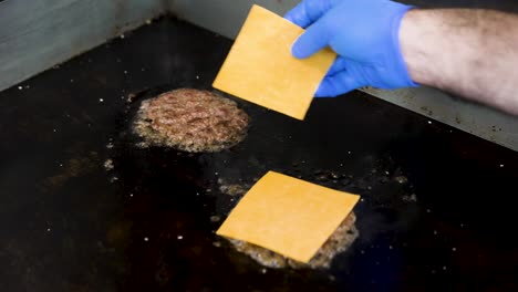 two burger patties sizzle on a grill as a chef places a slice of american cheese on each to create a smash cheeseburger