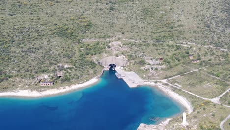 Vista-De-Drones-En-Albania-Dando-Vueltas-Frente-A-Un-Búnker-Naval-Con-Un-Barco-Como-Una-Cueva-En-Una-Montaña-Verde-Y-Un-Océano-Azul-En-Un-Día-Soleado