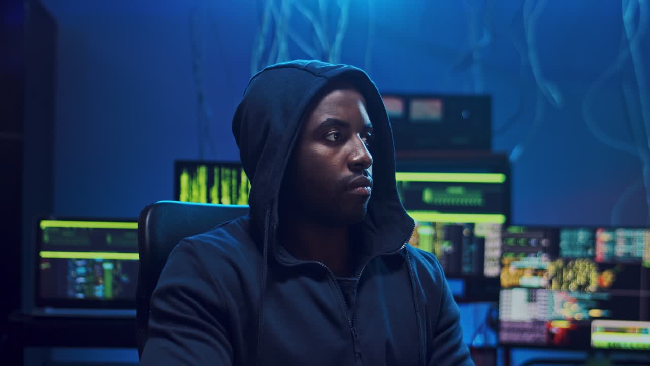 Portrait Shot Of The Young In A Hood Sitting In The Dark Room With Computers, Turning Face To The Camera Wth A Smile