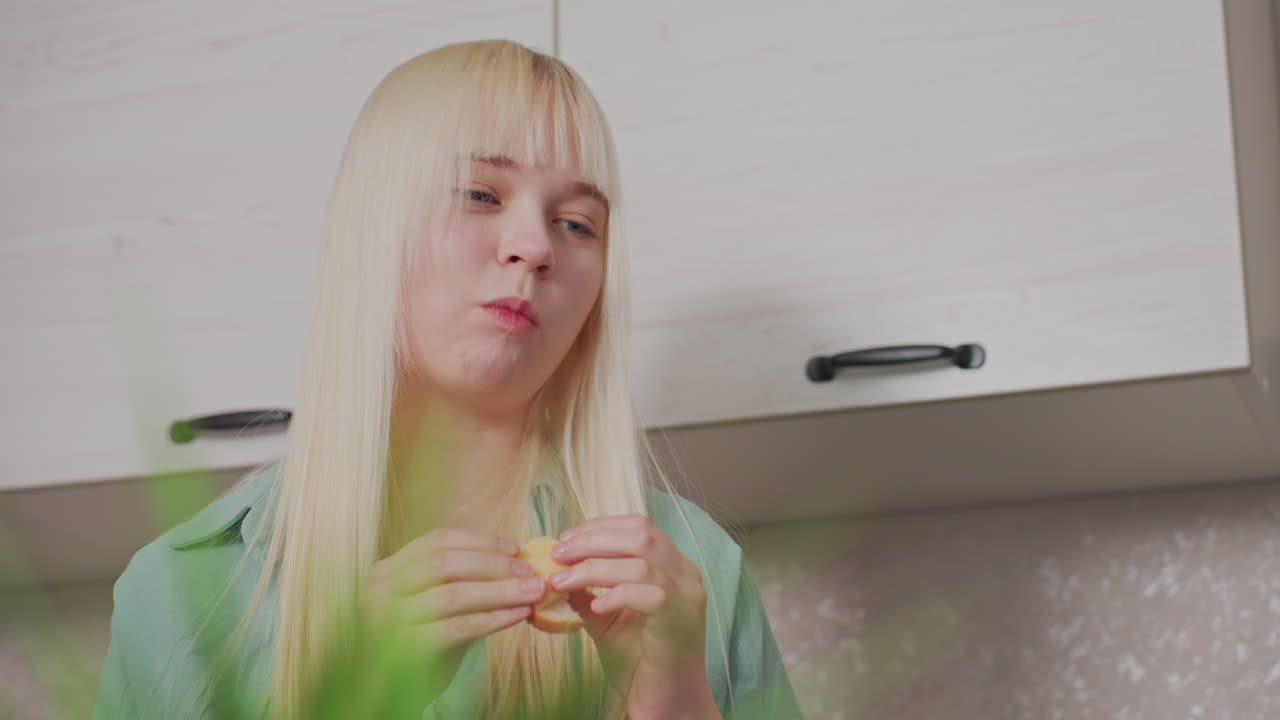 blonde girl in green shirt holding sandwich with both hands, smiling and taking bite while standing in modern kitchen with light-colored cabinets behind her