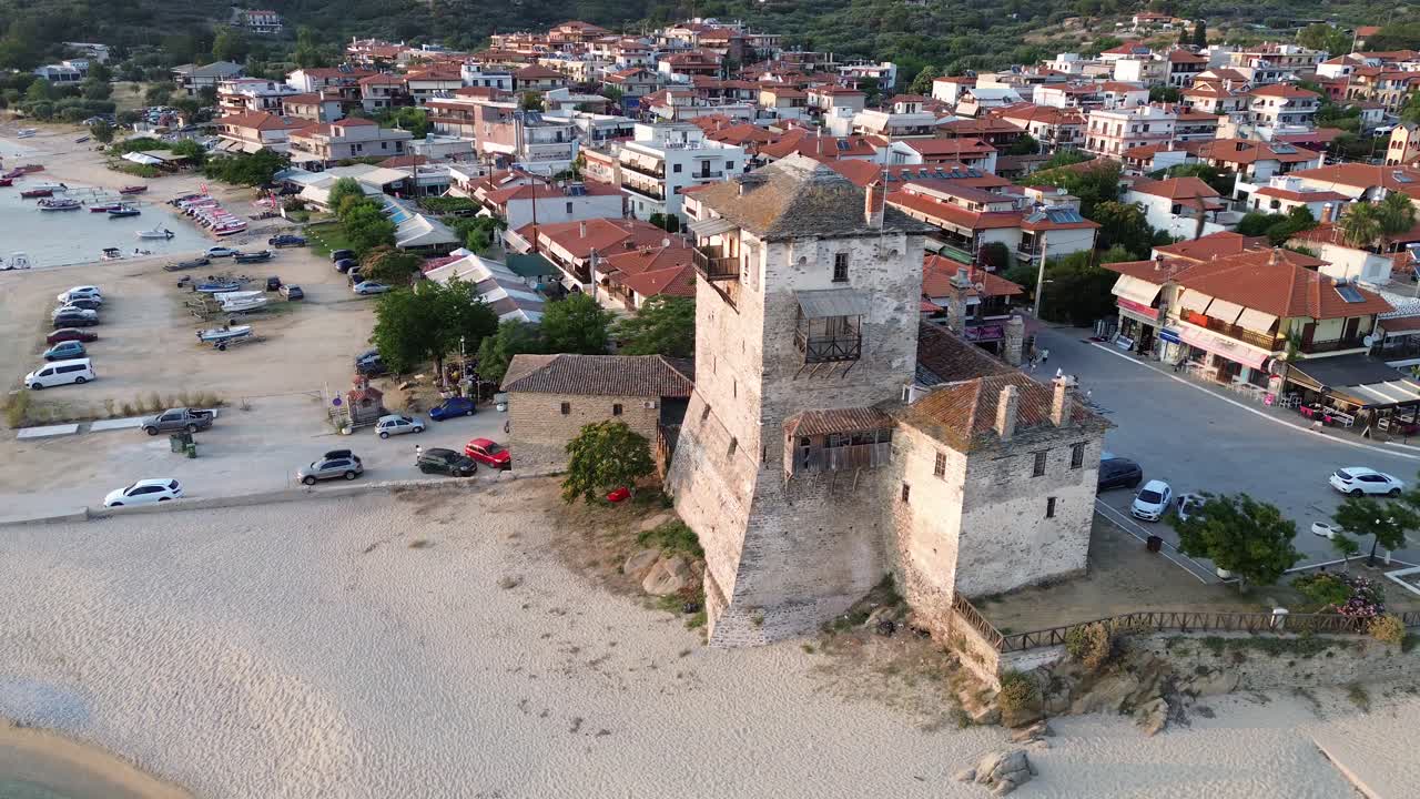 Orbita cinematografica di un drone intorno alla storica Torre di Ouranoupoli al tramonto, che mostra la struttura medievale in pietra in riva al mare e la porta del Monte Athos