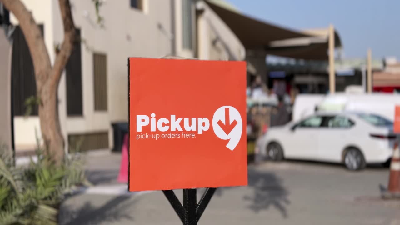 Primer plano de un letrero naranja de recogida de pedidos en un camino comercial exterior durante el día, con coche estacionado, pasarela y poca profundidad de campo en el fondo.