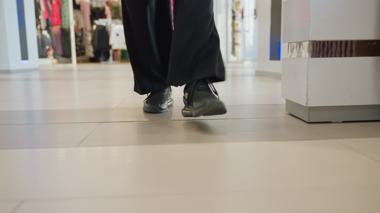 Low angle view of person walking indoors wearing black trousers and dark sneakers on polished tile floor near wooden bench and glowing store lights, background shows vibrant clothing display
