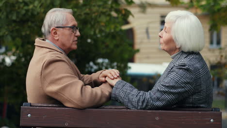 side view of elderly married couple holding hand and talking sitting on the bench in the park in autumn