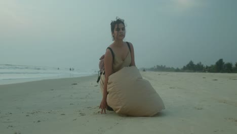a woman in a light dress, with her hair in a bun, kneels on a serene beach at dusk, drawing in the sand with her finger