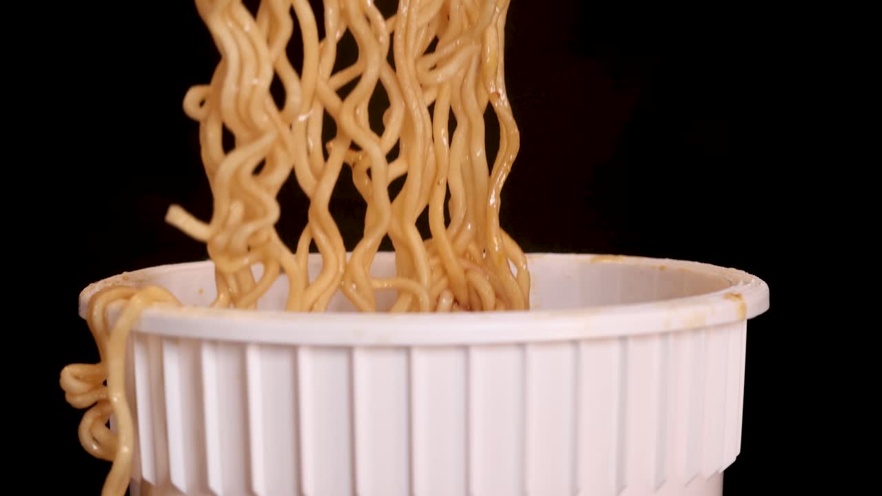 Hand lifts steaming instant noodles from white cup with chopsticks, close-up on dark background